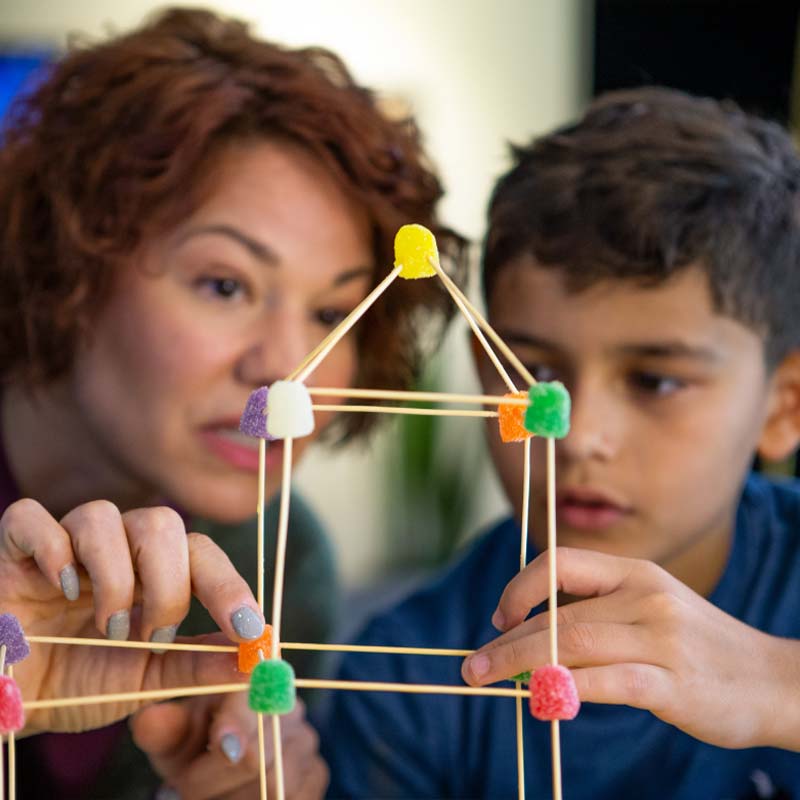 Adult woman helping a boy build a structure out of toothpicks and gumdrops.